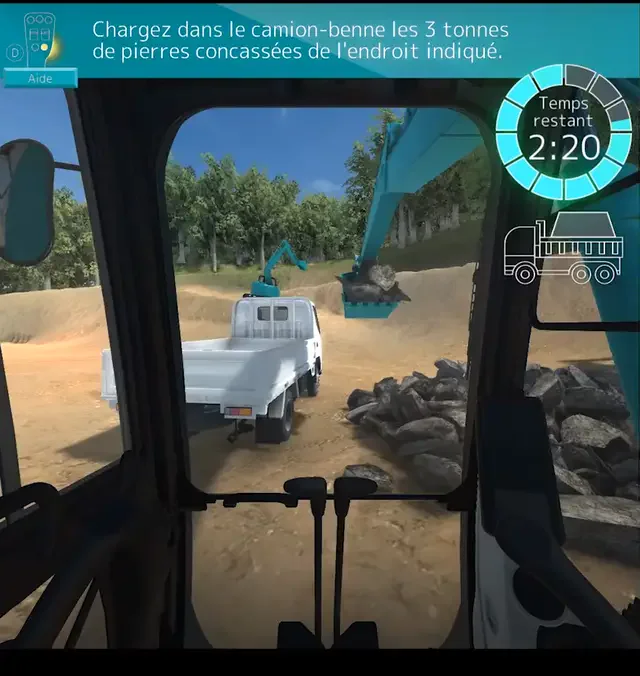ショベルの運転席から見た仮想空間の現場。画面の上にフランス語の課題の文、右上に残り時間の表示とダンプトラックの絵があり、ショベルが石をすくってトラックに積もうとしている